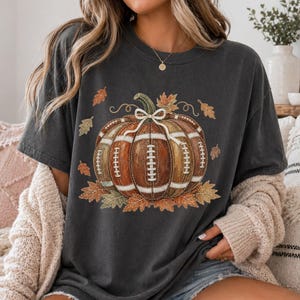 May include: A charcoal gray short-sleeve t-shirt featuring a graphic of a pumpkin designed with the texture and white laces of an American football. The pumpkin is accented with a white bow and surrounded by various autumn-colored fall leaves.