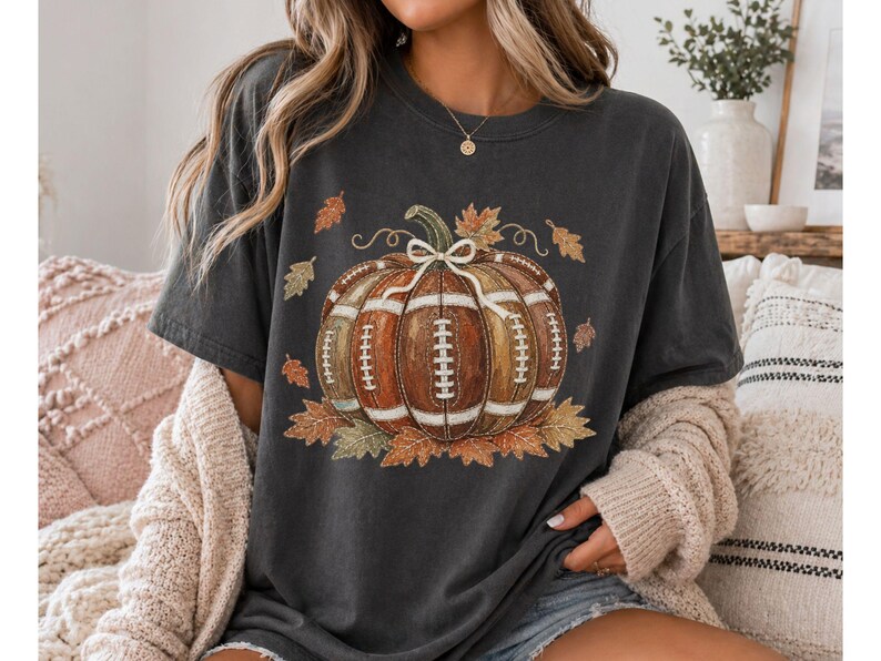 May include: A charcoal gray short-sleeve t-shirt featuring a graphic of a pumpkin designed with the texture and white laces of an American football. The pumpkin is accented with a white bow and surrounded by various autumn-colored fall leaves.