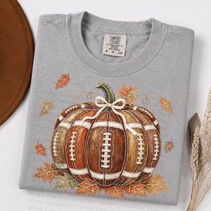 May include: A grey t-shirt featuring a graphic of a pumpkin designed with brown football-textured panels and white laces. The pumpkin is tied with a white string bow at the top and surrounded by scattered orange and yellow autumn leaves.