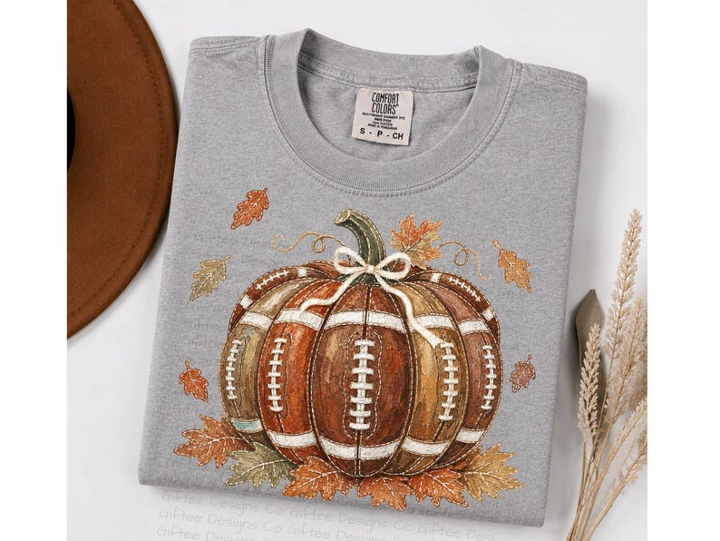 May include: A grey t-shirt featuring a graphic of a pumpkin designed with brown football-textured panels and white laces. The pumpkin is tied with a white string bow at the top and surrounded by scattered orange and yellow autumn leaves.