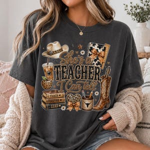 May include: A charcoal gray t-shirt featuring a graphic print with the text "She's a Teacher, I Can tell". The design includes a cowboy hat, a leopard print apple, a stack of books labeled "Teach, Inspire, Lead", a pencil holder, and a cowboy boot.