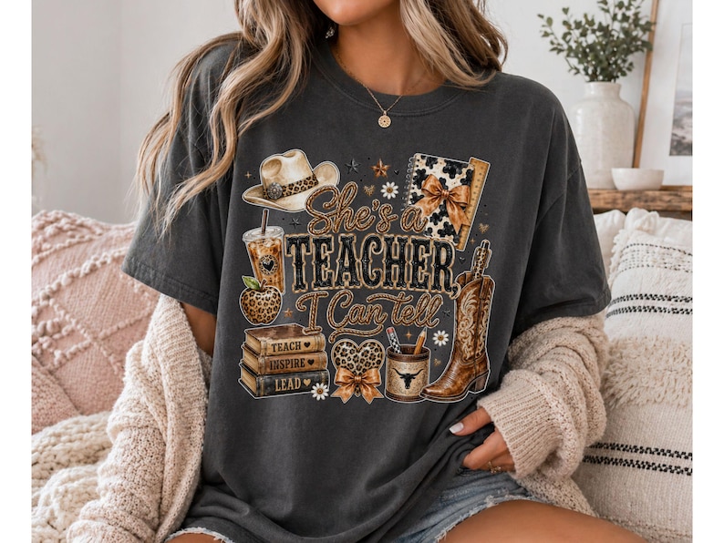 May include: A charcoal gray t-shirt featuring a graphic print with the text "She's a Teacher, I Can tell". The design includes a cowboy hat, a leopard print apple, a stack of books labeled "Teach, Inspire, Lead", a pencil holder, and a cowboy boot.