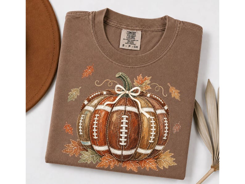 May include: A brown short-sleeved t-shirt featuring a graphic of a pumpkin designed with the texture and white laces of an American football. The design includes scattered autumn leaves and a small white bow tied around the pumpkin stem.