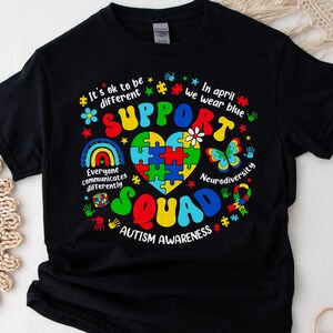 May include: Black t-shirt with a colorful puzzle piece heart design and the text "Support Squad Autism Awareness". The text "It's ok to be different" and "In April we wear blue" are also included.