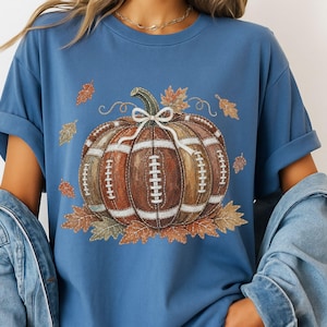 May include: A blue short-sleeve t-shirt featuring a central graphic of a pumpkin designed with the texture and white laces of an American football. The pumpkin is accented with a white bow and surrounded by various autumn-colored fall leaves.