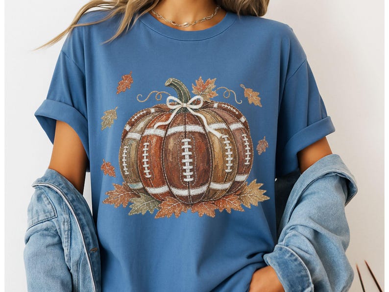 May include: A blue short-sleeve t-shirt featuring a central graphic of a pumpkin designed with the texture and white laces of an American football. The pumpkin is accented with a white bow and surrounded by various autumn-colored fall leaves.