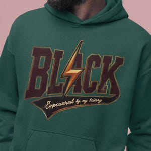 May include: A dark green hoodie with a vintage-style graphic design. The graphic features the word "BLACK" in large, bold letters with a lightning bolt in the center. The text "Empowered by my history" is written in a smaller font below the word "BLACK".