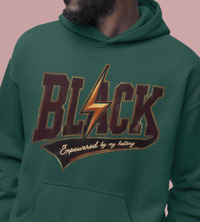 May include: A dark green hoodie with a vintage-style graphic design. The graphic features the word "BLACK" in large, bold letters with a lightning bolt in the center. The text "Empowered by my history" is written in a smaller font below the word "BLACK".