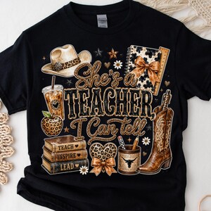 May include: A black t-shirt featuring a graphic print with the text "She's a TEACHER I Can tell". The design includes a cowboy hat, iced coffee, leopard print apple, stack of books labeled "TEACH", "INSPIRE", "LEAD", a heart, and a cowboy boot.