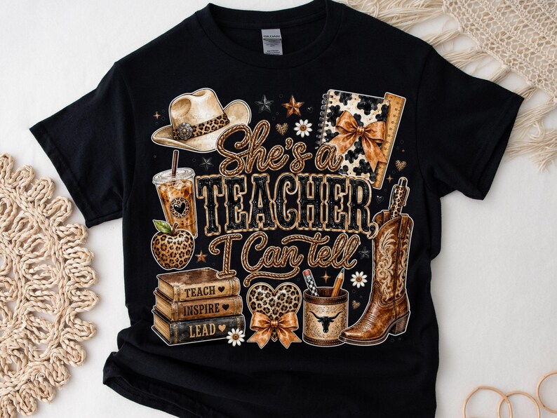 May include: A black t-shirt featuring a graphic print with the text "She's a TEACHER I Can tell". The design includes a cowboy hat, iced coffee, leopard print apple, stack of books labeled "TEACH", "INSPIRE", "LEAD", a heart, and a cowboy boot.