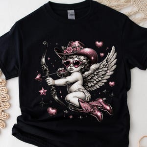 May include: Black t-shirt with a pink and white cherub wearing a cowboy hat and heart-shaped sunglasses holding a bow and arrow. The cherub has wings and is surrounded by hearts and stars.