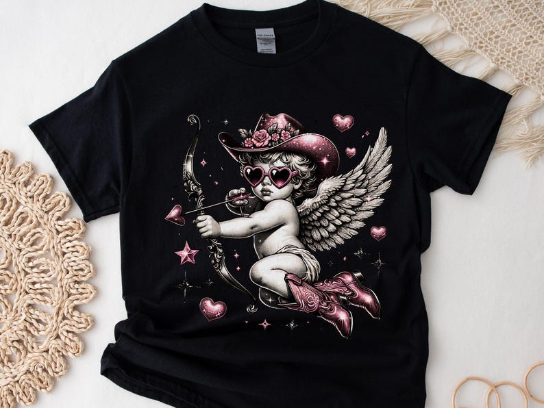 May include: Black t-shirt with a pink and white cherub wearing a cowboy hat and heart-shaped sunglasses holding a bow and arrow. The cherub has wings and is surrounded by hearts and stars.