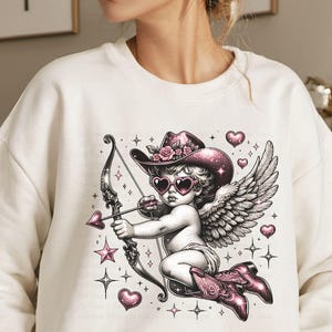 May include: White sweatshirt with a graphic print of a cupid with pink wings, a pink hat with flowers, and pink sunglasses holding a bow and arrow. The cupid is surrounded by pink hearts and stars.