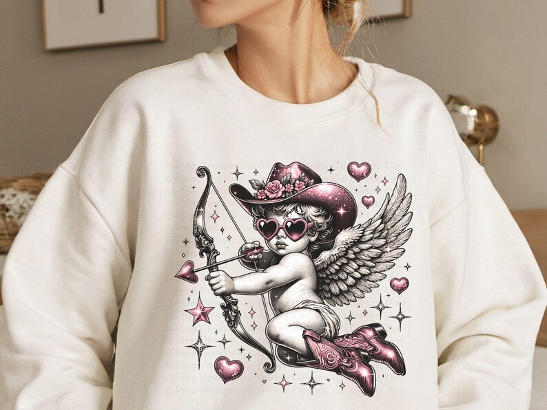 May include: White sweatshirt with a graphic print of a cupid with pink wings, a pink hat with flowers, and pink sunglasses holding a bow and arrow. The cupid is surrounded by pink hearts and stars.