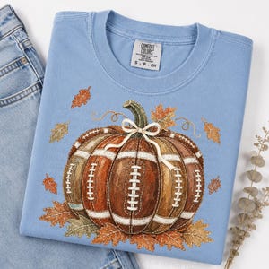 May include: A light blue crew neck t-shirt featuring a graphic of a pumpkin designed with the texture and white laces of an American football. The pumpkin is accented with a small white bow and surrounded by various orange and brown autumn leaves.