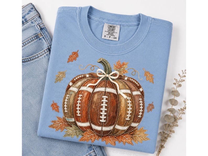 May include: A light blue crew neck t-shirt featuring a graphic of a pumpkin designed with the texture and white laces of an American football. The pumpkin is accented with a small white bow and surrounded by various orange and brown autumn leaves.