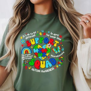 May include: Green t-shirt with a colorful graphic design that says "Support Squad" and includes a puzzle piece heart, rainbows, butterflies, and the text "It's ok to be different", "Everyone communicates differently", "In April we wear blue", "Neurodiversity", and "Autism Awareness".