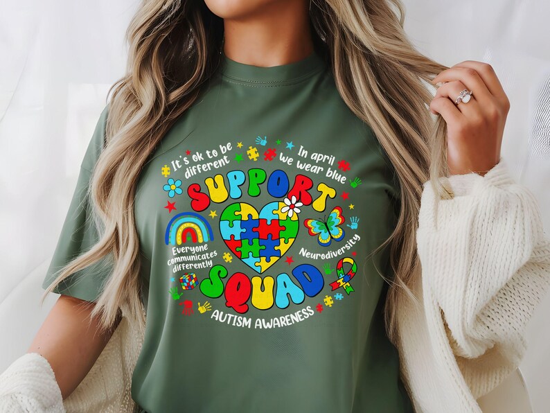 May include: Green t-shirt with a colorful graphic design that says "Support Squad" and includes a puzzle piece heart, rainbows, butterflies, and the text "It's ok to be different", "Everyone communicates differently", "In April we wear blue", "Neurodiversity", and "Autism Awareness".