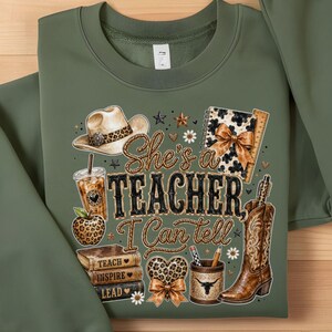 May include: An olive green crewneck sweatshirt featuring a graphic print with the text "She's a TEACHER, I Can tell". The design includes a cowboy hat, a leopard print apple, a stack of books, a pencil holder, a cowboy boot, and a notebook.