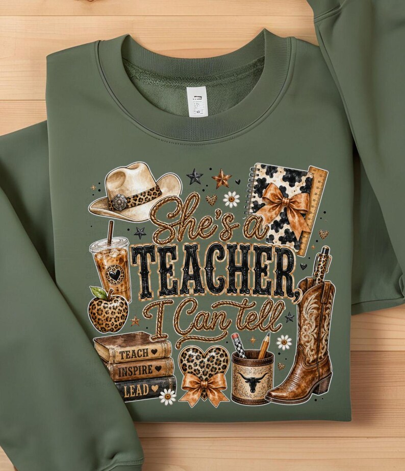 May include: An olive green crewneck sweatshirt featuring a graphic print with the text "She's a TEACHER, I Can tell". The design includes a cowboy hat, a leopard print apple, a stack of books, a pencil holder, a cowboy boot, and a notebook.