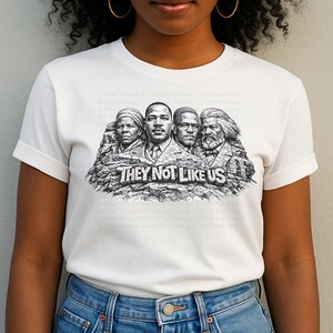 以下が含まれることがあります： 「THEY NOT LIKE US」の文字と、岩に彫られた4人の歴史上の人物の白黒グラフィックが特徴の白いTシャツ。青いジーンズを履いた人が着ています。