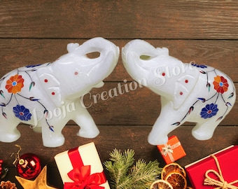 Puede incluir: Dos elefantes de mármol blanco que se miran, decorados con coloridos patrones florales y detalles intrincados. Los elefantes están sobre una superficie de madera con decoraciones navideñas.