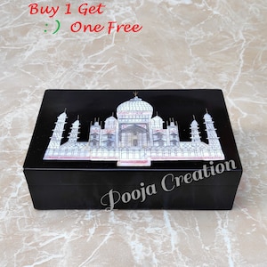 May include: Black wooden box with a white inlay of the Taj Mahal. The box has the words "Pooja Creation" on the front.