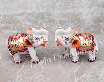 Puede incluir: Dos figuritas de elefante de mármol blanco con detalles pintados en rojo, dorado y verde. Los elefantes se miran y tienen un pequeño colmillo pintado de naranja a cada lado. Los elefantes están decorados con diamantes de imitación.