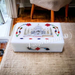 May include: White marble box with a colorful inlay design of the Taj Mahal. The box is decorated with red, blue, green, and gold accents. The text "Pooja Creati(v)" is visible on the bottom of the box.