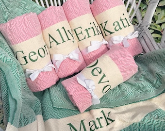 May include: Pink and cream-colored personalized towels with names in green lettering, tied with white ribbons. The towels are arranged on a green and white patterned blanket, creating a coordinated set.