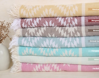 May include: A stack of seven patterned towels in various colors, including mustard, burgundy, brown, petrol green, turquoise, mint, and fuchsia. The towels have a white fringe and a geometric pattern.