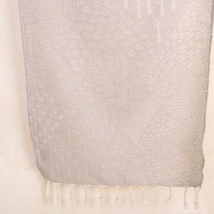 May include: A light gray scarf with a detailed, textured pattern. The design includes geometric shapes and vertical lines, with a fringe at the bottom. The scarf is made of a soft material and is suitable for various occasions.