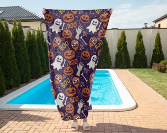 Può includere: Un asciugamano a tema Halloween con un motivo ripetuto di zucche arancioni, fantasmi bianchi, ossa e la parola "BOO" su uno sfondo viola scuro. L'asciugamano è tenuto davanti a una piscina.