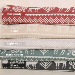 May include: A stack of folded blankets in burgundy, beige, light gray, dark green, and black. Each blanket features a repeating pattern of deer and geometric shapes. The edges have cream-colored fringe, and the color of each blanket is labeled.