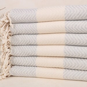 May include: A stack of six folded, light grey and cream-coloured herringbone-patterned towels. The towels have a fringed edge and are stacked neatly. The towels are made of a soft, absorbent material, ideal for use in a bathroom or spa.