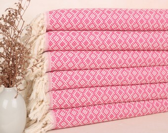Può includere: Una pila di asciugamani rosa e bianchi a fantasia con bordi sfrangiati. Gli asciugamani hanno un design geometrico e sono ben piegati. Un vaso bianco con fiori secchi è sullo sfondo.