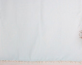 May include: A pale blue beach towel with a textured weave and a fringe along the bottom edge. The towel is displayed against a neutral background, with a vase of dried flowers to the right.