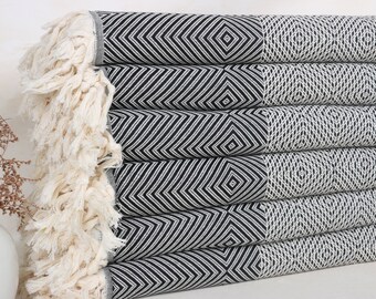 May include: A pile of black and white patterned towels with a geometric design. The towels feature a diamond and chevron pattern. The edges have a cream-coloured fringe. The towels are folded and stacked neatly. These towels are suitable for a bathroom or spa.