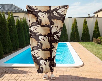 Può includere: Una coperta a tema Halloween con un motivo ripetuto di fantasmi dei cartoni animati nei toni del beige e del bianco su uno sfondo marrone scuro. La coperta è tenuta davanti a una piscina e alberi verdi.
