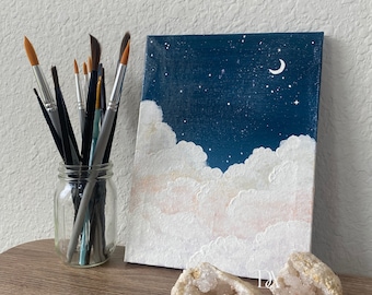 May include: A painting of a night sky with white clouds and a crescent moon. The painting is on a canvas and is propped up against a white wall. There are paintbrushes in a glass jar in the foreground.