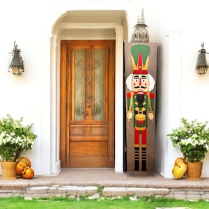 May include: A large, wooden nutcracker decoration stands beside a wooden door. The nutcracker is painted with a green and red uniform, a white beard, and a tall green hat. The door is framed by white walls and flanked by potted plants.