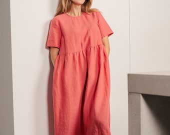 Puede incluir: Una mujer lleva un vestido de lino coral con mangas cortas y un corte holgado. El vestido tiene una cintura fruncida y le llega a las rodillas. Está de pie frente a una pared blanca.