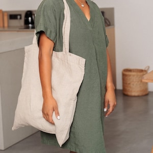 Könnte beinhalten: Eine Frau in einem grünen Leinenkleid und einer silbernen Halskette hält eine beige Tragetasche.