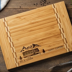 May include: A rectangular bamboo cutting board with a camper design and the text "Happy Campers" and "Bill & Joanna". Kitchen utensils, including a knife, wooden spoon, and spatula, are arranged around the board.