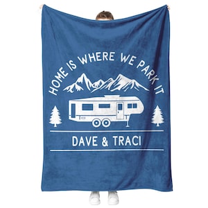 May include: A blue blanket with white text and graphics. The text reads "HOME IS WHERE WE PARK IT" above a drawing of a camper and mountains. Below the camper is the text "DAVE & TRACI".