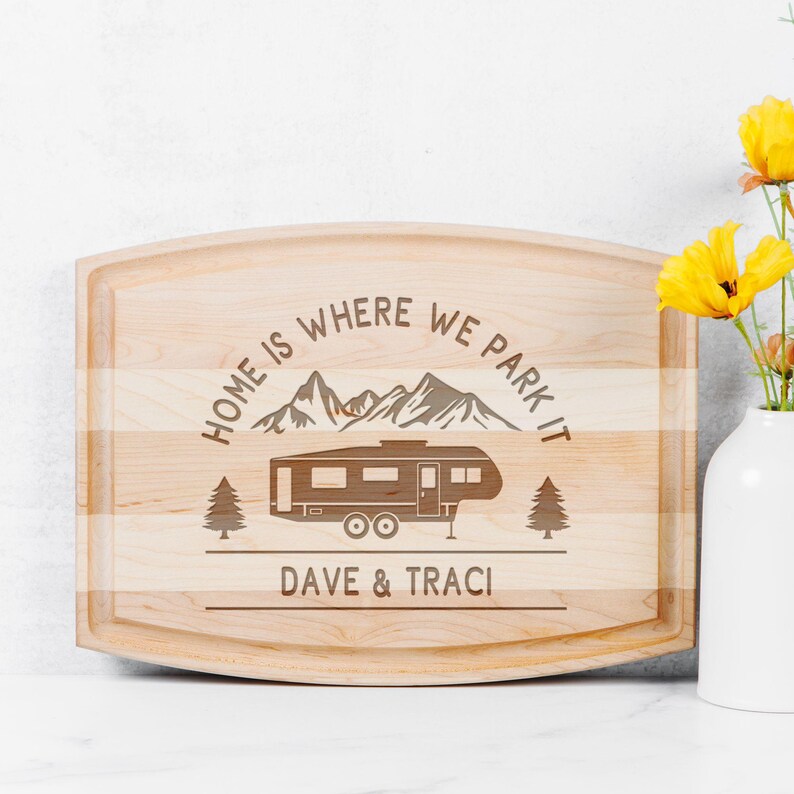May include: A light-colored wooden cutting board with rounded edges. The board features an engraved design of a camper, mountains, and trees, with the text "HOME IS WHERE WE PARK IT" and the names "DAVE & TRACI".