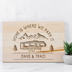 May include: A light-colored wooden sign with the phrase "HOME IS WHERE WE PARK IT" above a camper illustration. The sign also includes the names "DAVE & TRACI" below the camper, and two trees on either side.