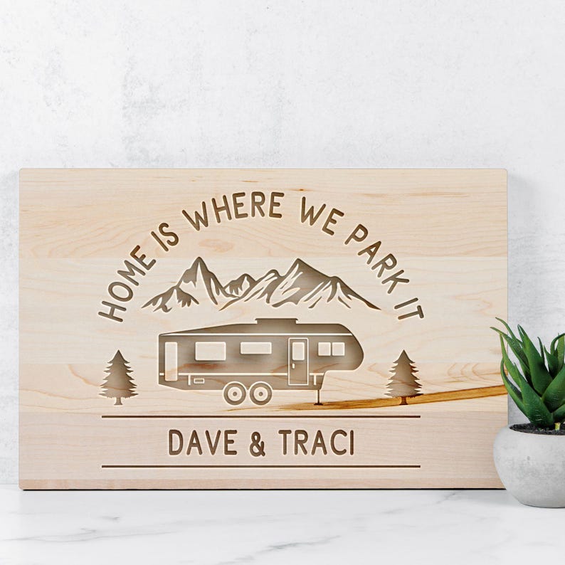 May include: A light-colored wooden sign with the phrase "HOME IS WHERE WE PARK IT" above a camper illustration. The sign also includes the names "DAVE & TRACI" below the camper, and two trees on either side.