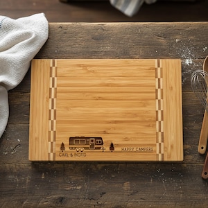 May include: A rectangular bamboo cutting board with a camper design and the words "Happy Campers" and "Carl & Ingrid" engraved on it. A white towel and cooking utensils are next to the board.