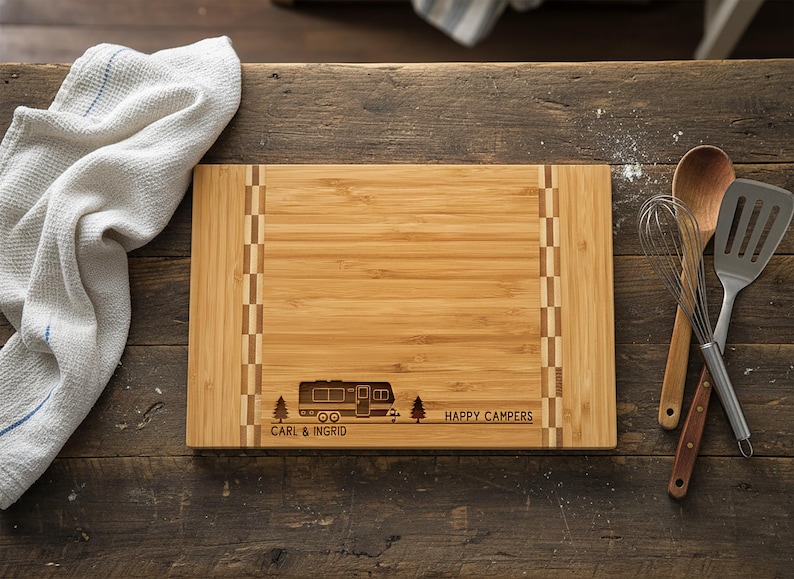 May include: A rectangular bamboo cutting board with a camper design and the words "Happy Campers" and "Carl & Ingrid" engraved on it. A white towel and cooking utensils are next to the board.